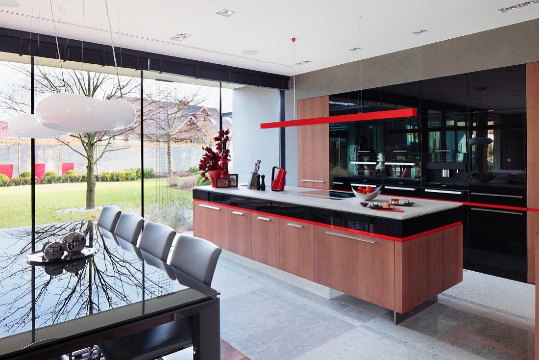 Teilweise sichtbarer Essplatz vor Kochbereich mit Küchenblock unter roter, filigraner Hängeleuchte und Einbauschrank mit schwarzer Front, seitlich Glasfassade mit Blick in Innenhof