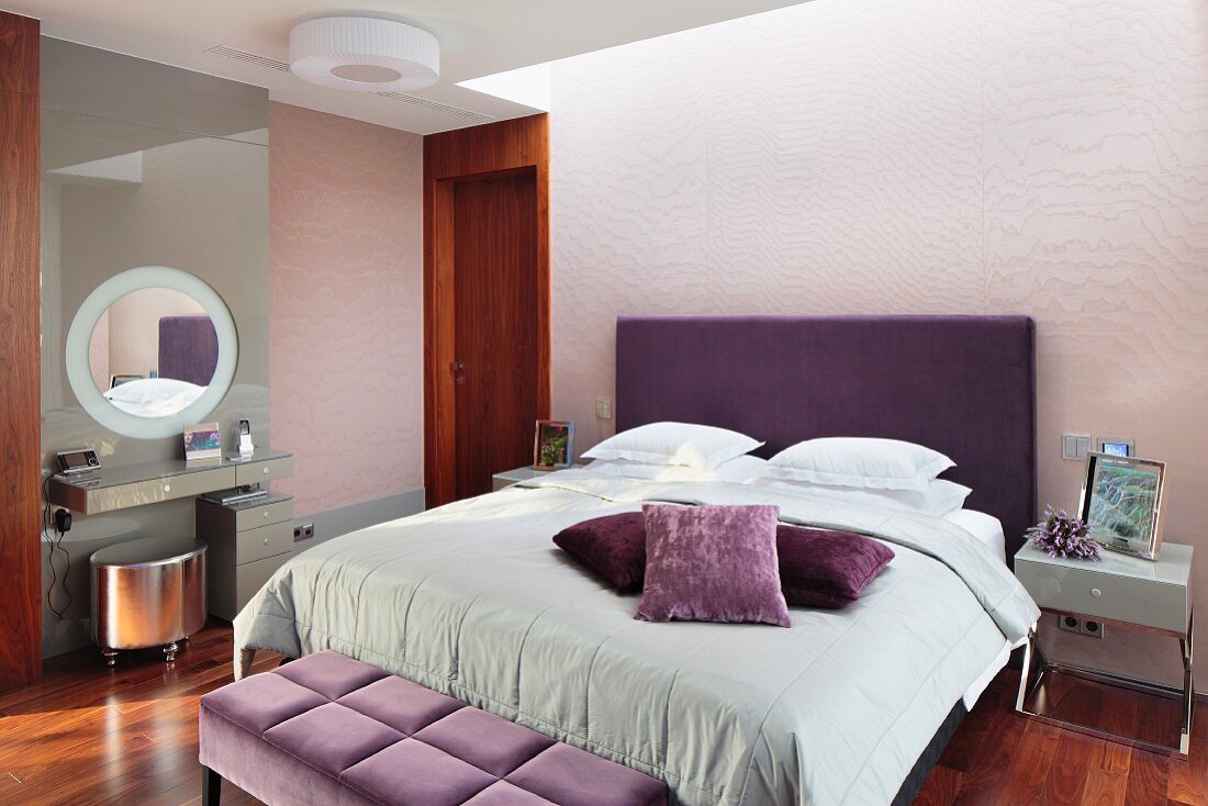 Elegant bedroom; double bed with purple upholstered headboard, scatter cushions in shades of purple on pale grey blanket, minimalist dressing table and silver-coloured stool
