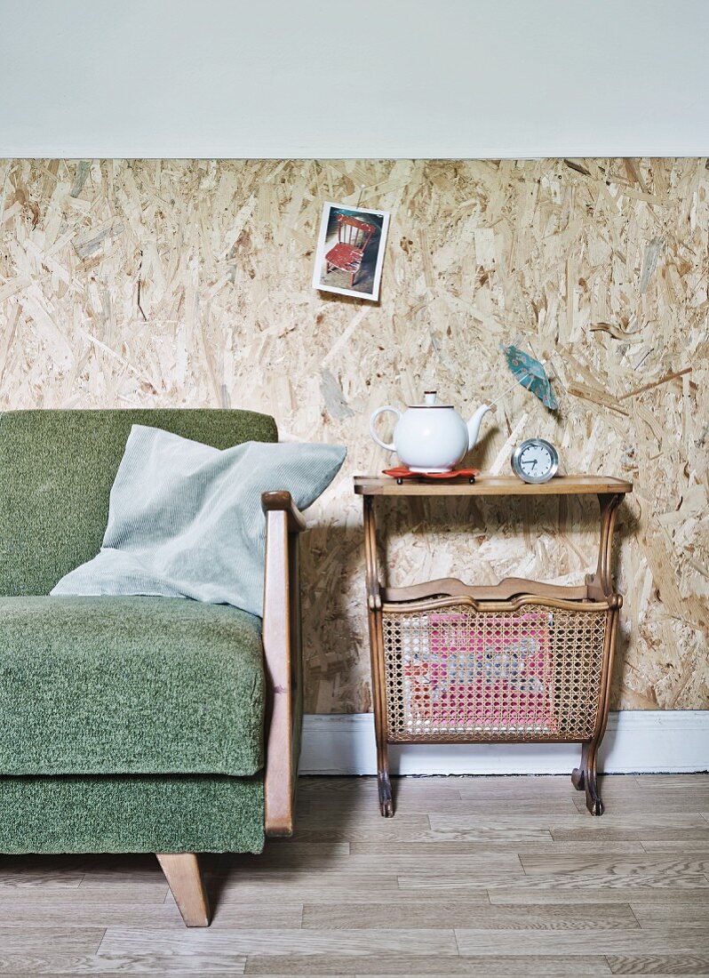 50s sofa and second-hand retro side table with magazine rack against wall clad in rustic chipboard panels