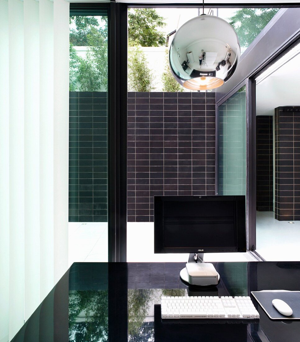 Monitor on a black table top in front of a room height bank of windows with a view of a patio with a black tiled wall