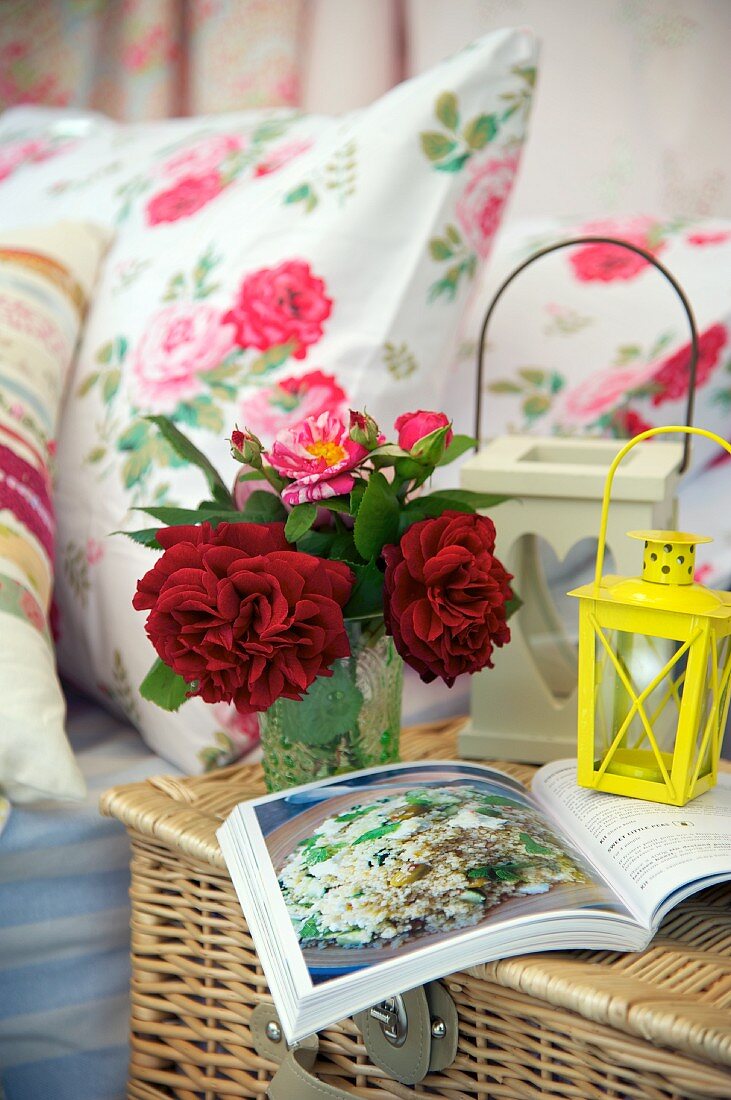 A small bunch of flowers, lanterns and a book on top of a picnic basket next to bed