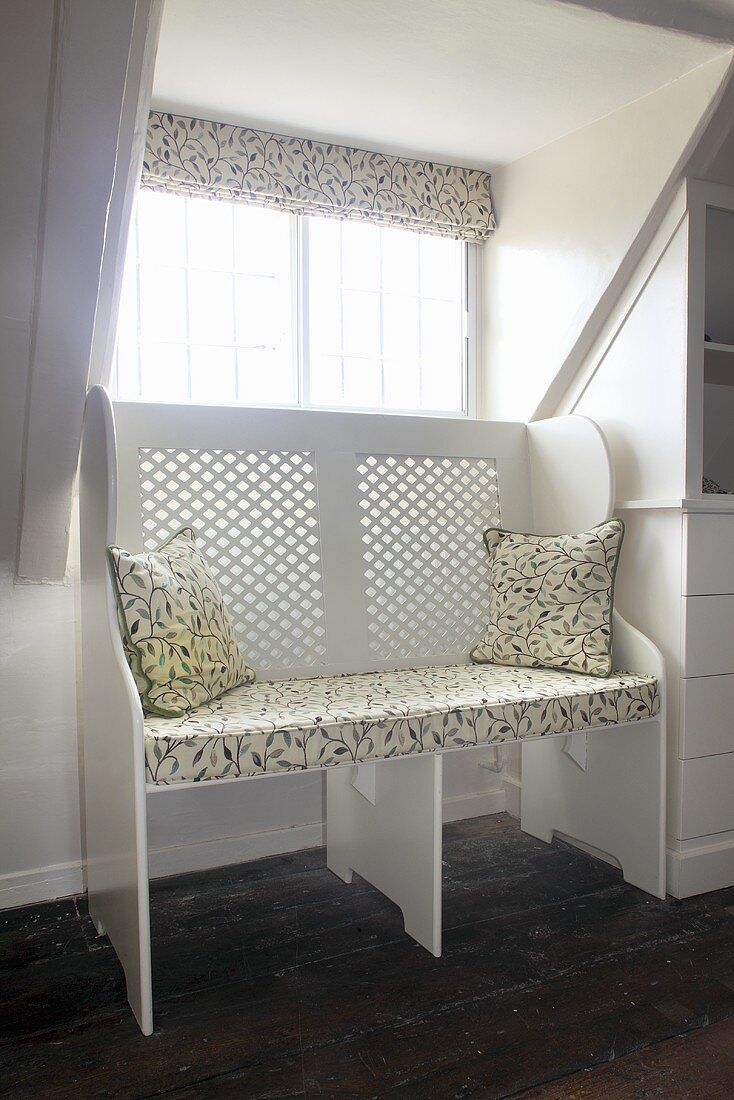 Eine weiße Sitzbank mit gemusterter Auflage und Dekokissen vor dem Fenster