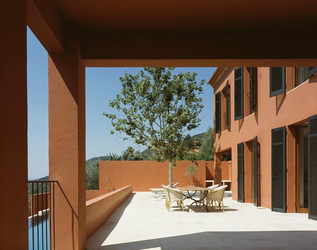 Sonnenbeschienene Terrasse mit Tisch und Stühlen vor Baum und mediterranem Haus mit rotbrauner Fassade