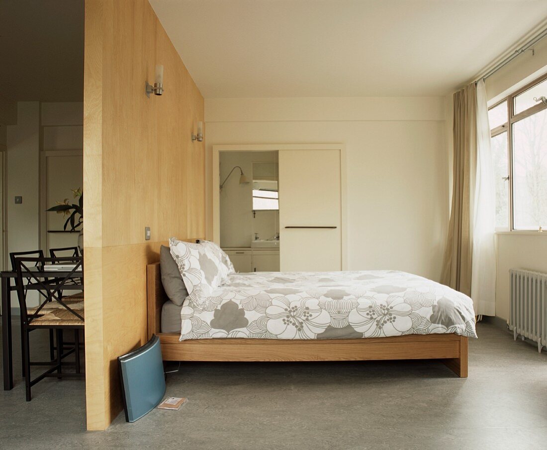 Modernes Bett mit Holzgestell vor Raumteiler aus Holz und Blick ins Bad ensuite und auf Stühle hinter Trennwand