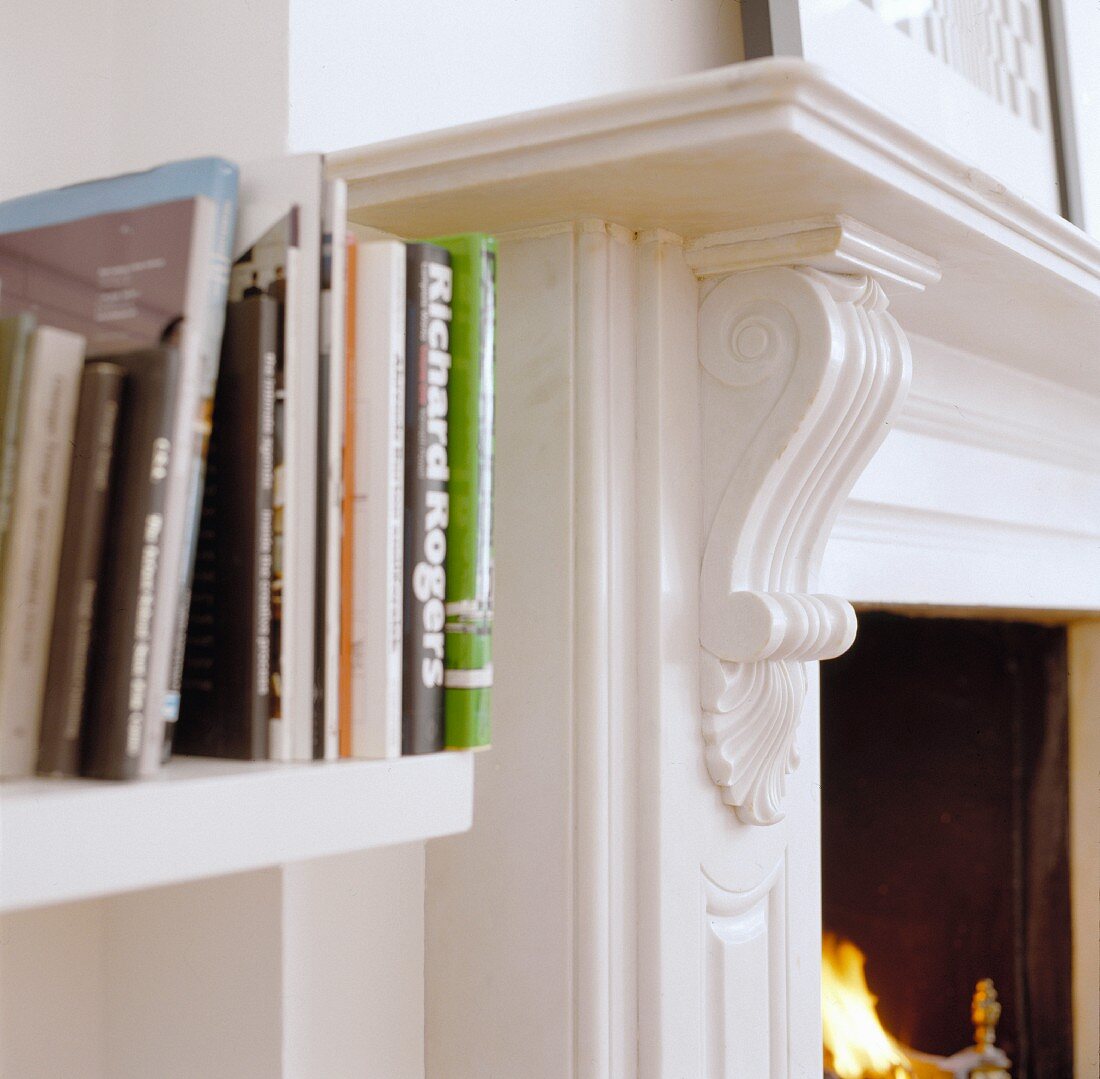 Detail of white shelves with books beside a chimney with a carved, white lacquered mantelpiece