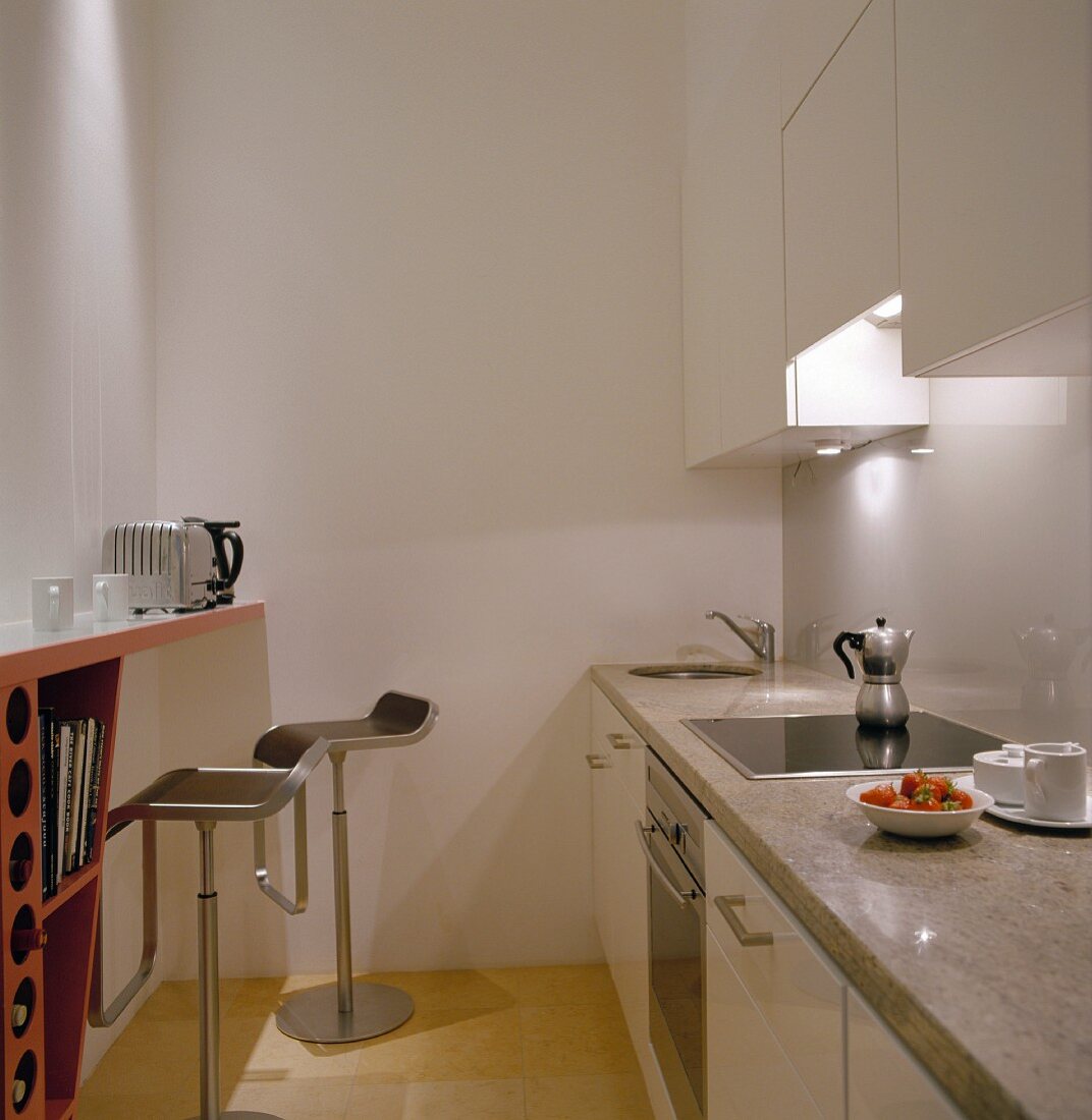 Küchenzeile mit weissen Fronten und Steinplatte gegenüber eingebauter Esstheke an Wand und Barhockern aus Edelstahl