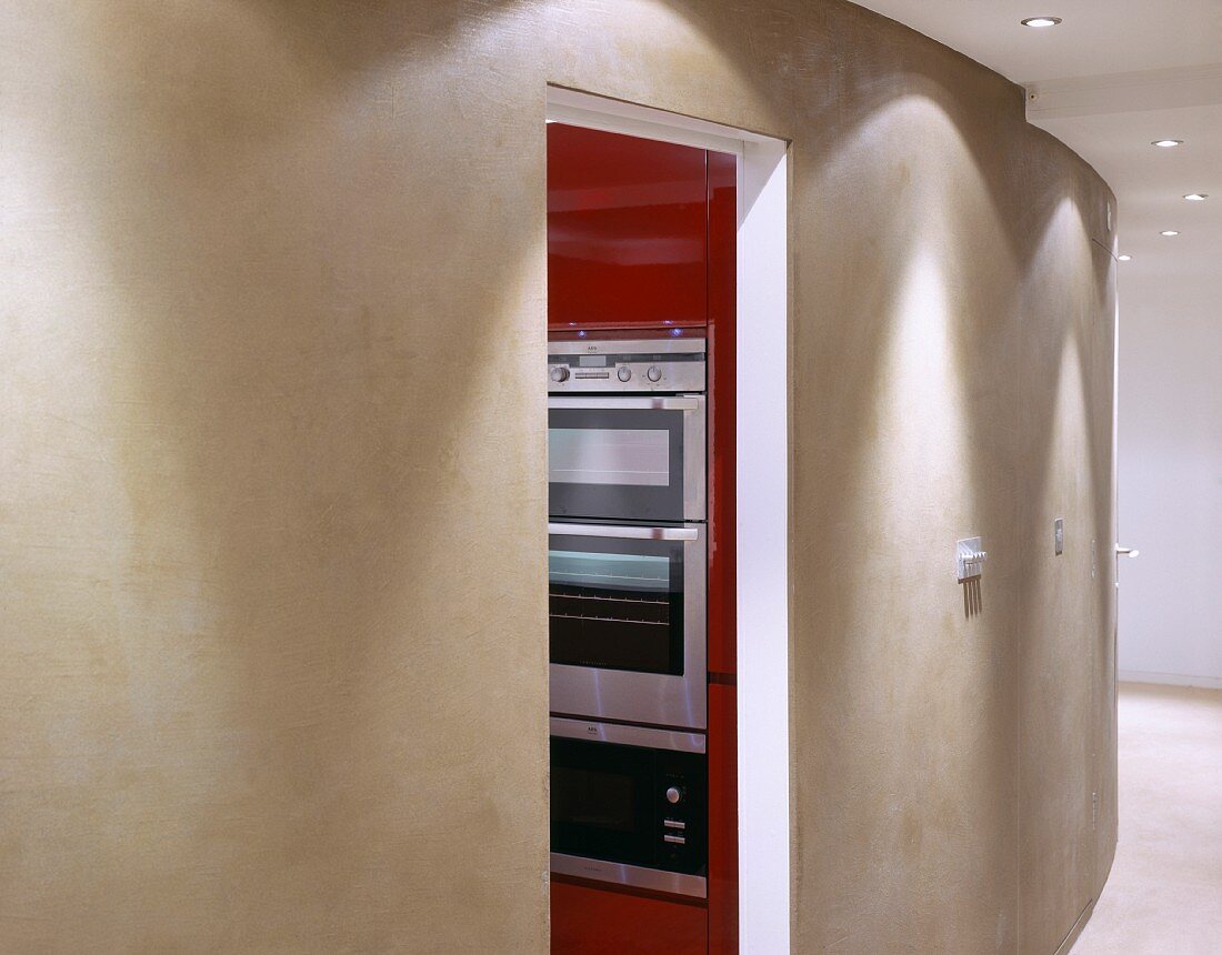 Raum mit geschwungener grau getönter Wand und offener Tür mit Blick auf Kücheneinbaugeräten