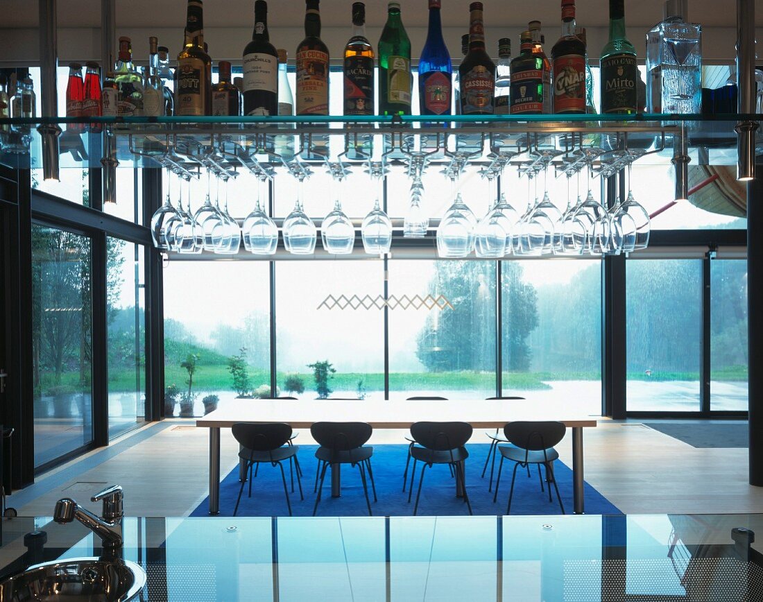 Bar mit Spirituosen und Gläsern auf abgehängtem Regal und Blick auf Esstisch vor Glasfassade