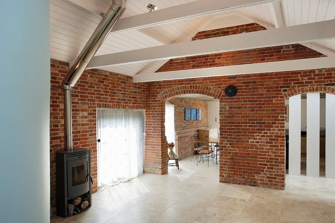 Living room in a converted barn with brick wall and openings with round arches under a white lacquered wood ceiling an wood burning stove