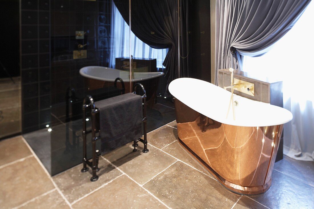 Freestanding bathtub clad in copper colored material in front of black, glossy cabinet on a tiled floor