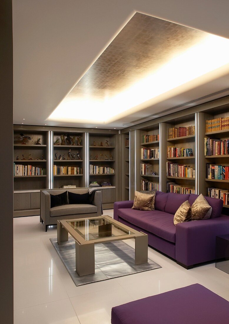 Wohnraum mit violettem Sofa vor Bücherwand und indirektem Licht unter offener Decke