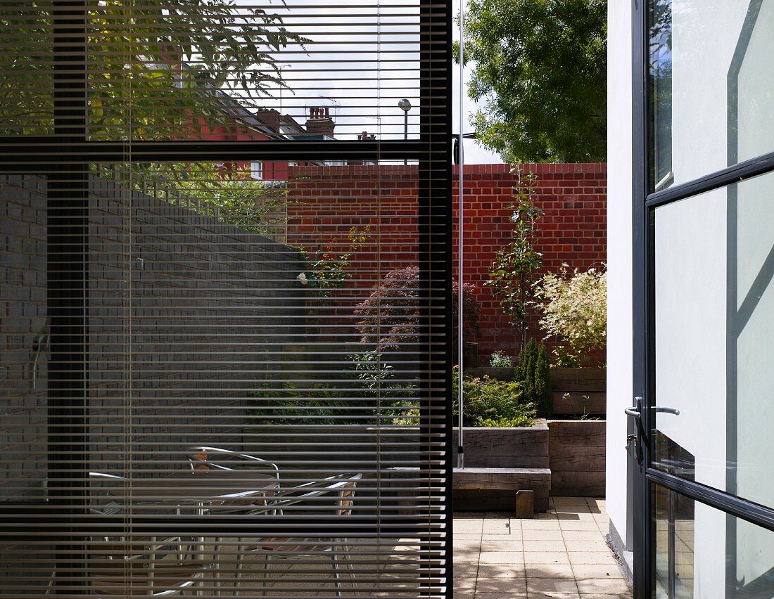Window with partially closed blinds and view through and open patio door of a city garden