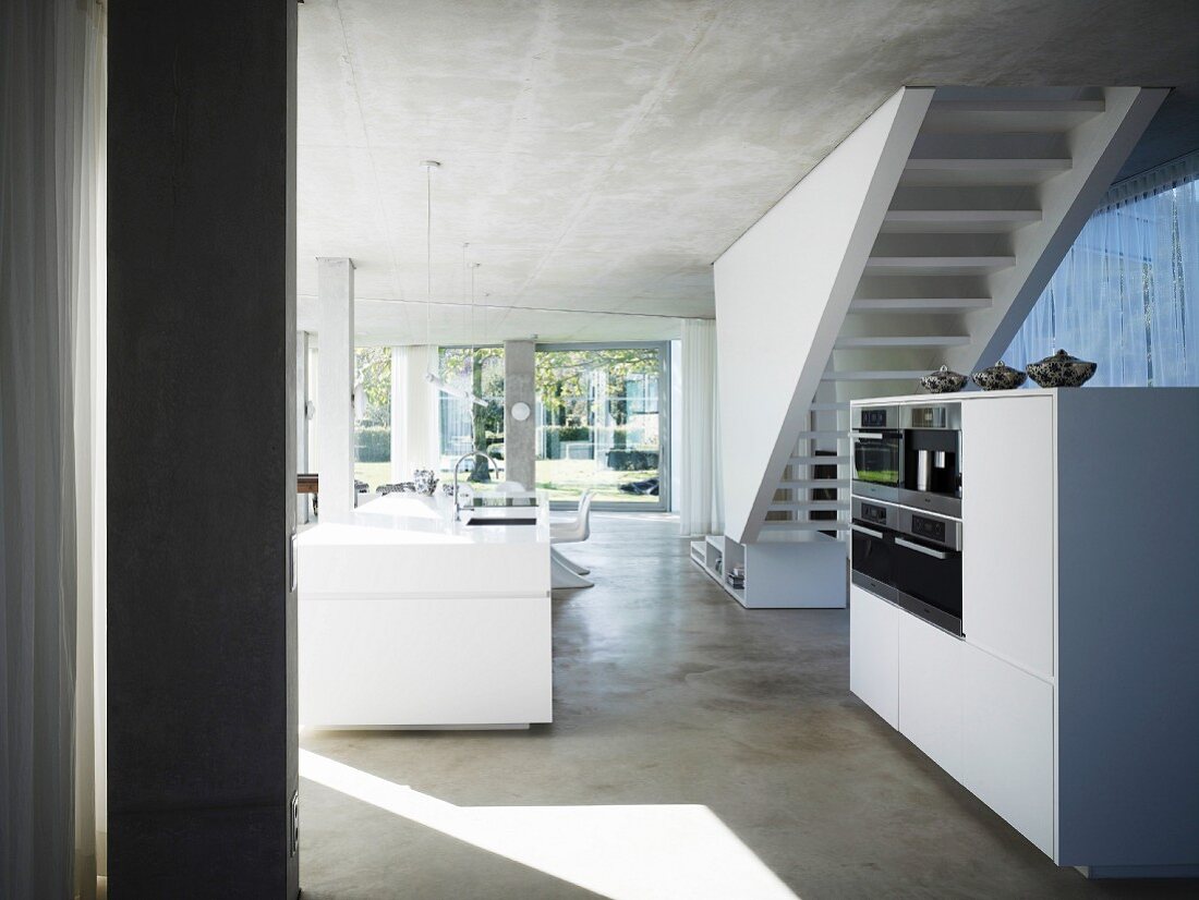 Open living room with concrete ceiling and floor and white free standing kitchen unit in front of a stairway