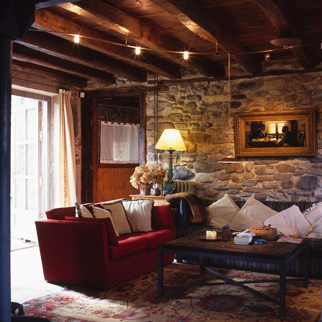 Old, living room in a country home with red couch and a sofa decorated with pillows in front of a natural stone wall