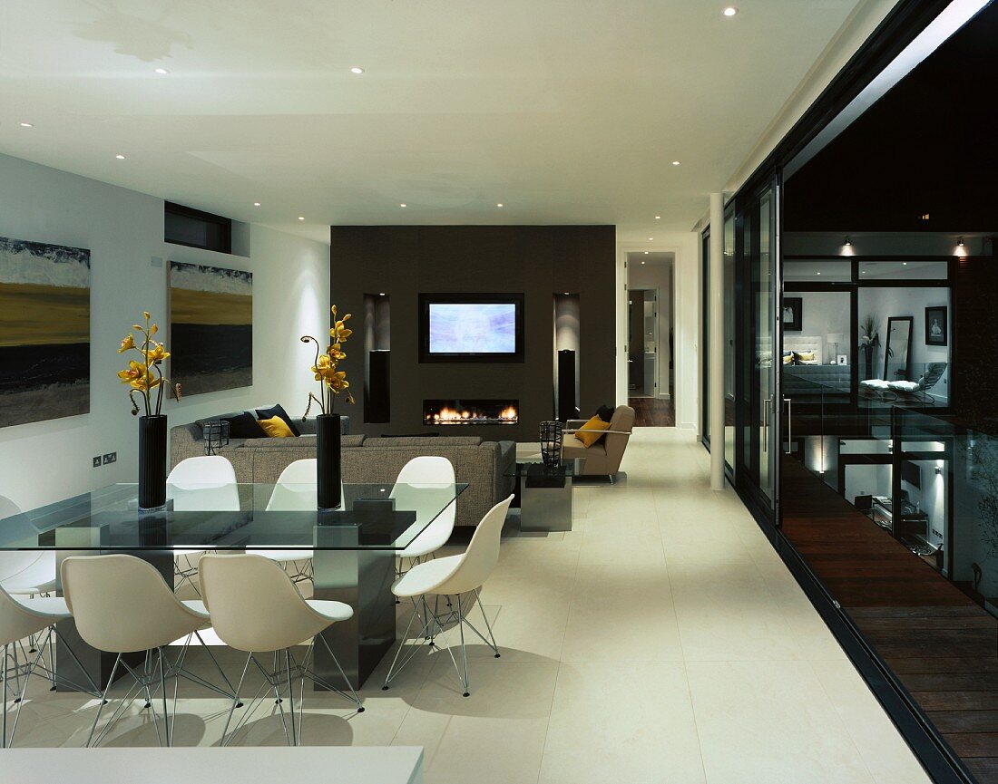 weiße Schalenstühle vor modernem Esstisch im offenen Wohnraum und offenstehender Terrassentür mit Blick auf Pool in Nachtstimmung