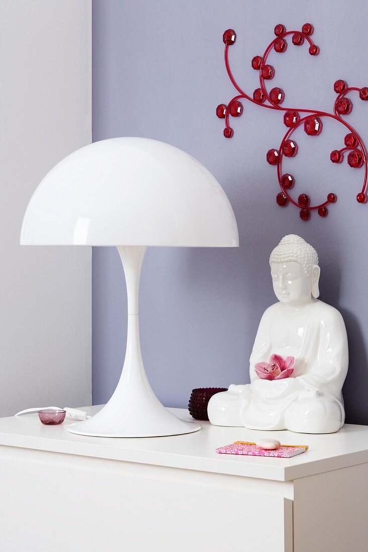 weiße Tischleuchte (Louis Poulsen/Verner Panton) mit Buddha Figur auf einer Kommode vor Wandobjekt