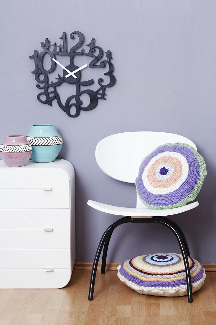 A wall clock above a chest of drawers and a chair with a cushion