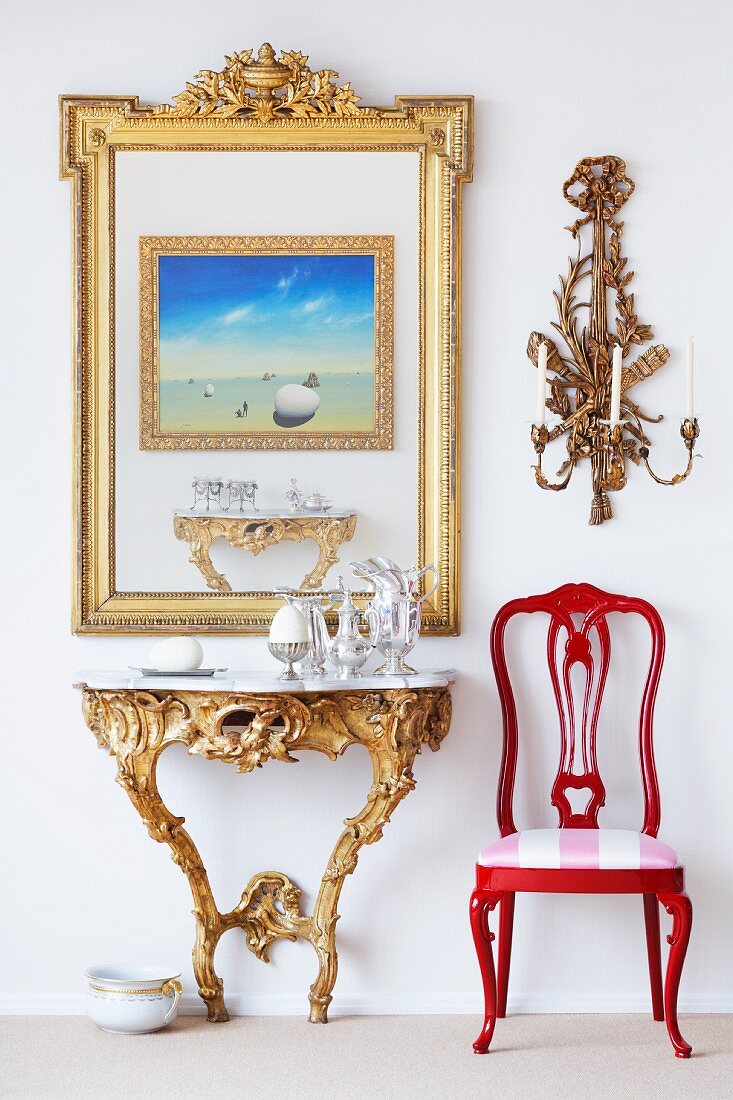 Wandtisch im Rokoko-Stil und ein roter Stuhl, darüber goldgerahmtes Bild und Wandkerzenhalter