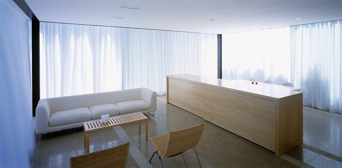 Moderner Wohnraum mit Sofa und monolithischer Theke aus Holz vor weissen geschlossenen Vorhängen am Fenster
