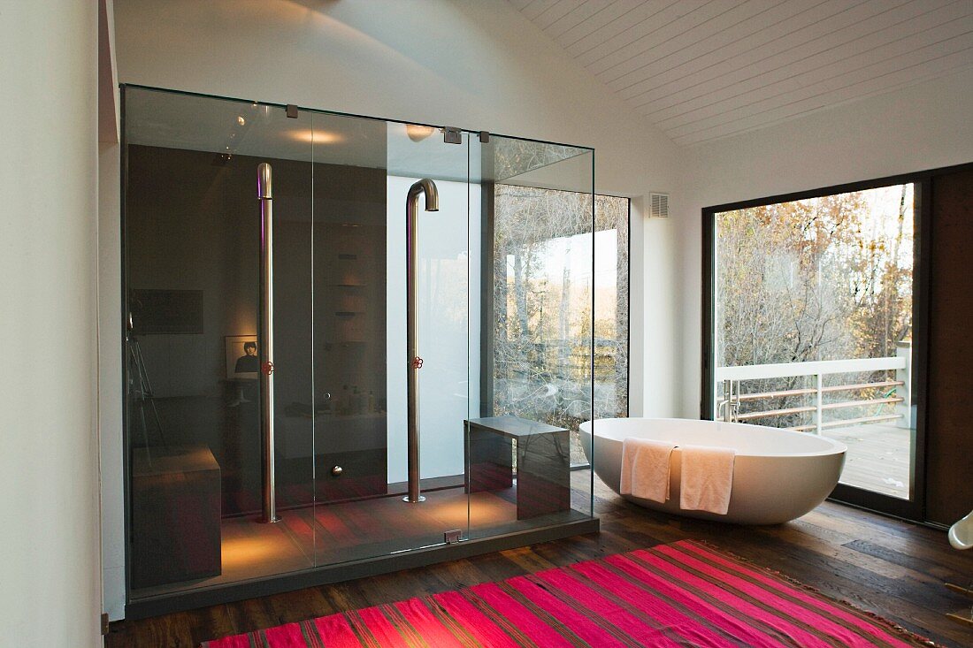 Freestanding bathtub in front of terrace windows next to a spacious glass shower stall with pink striped rug