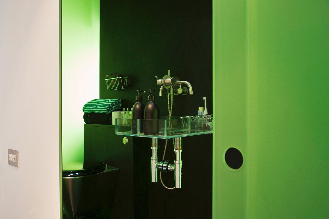 Blick ins offene Bad auf Ablage mit Badutensilien und grün gefärbte Glaswand