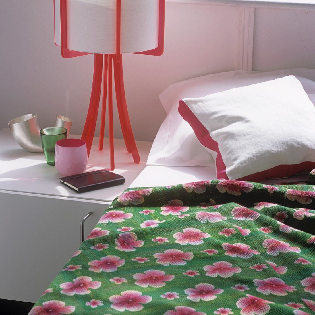 A bed with a floral patterned quilt and a table lamp with a red plastic base on a bedside table