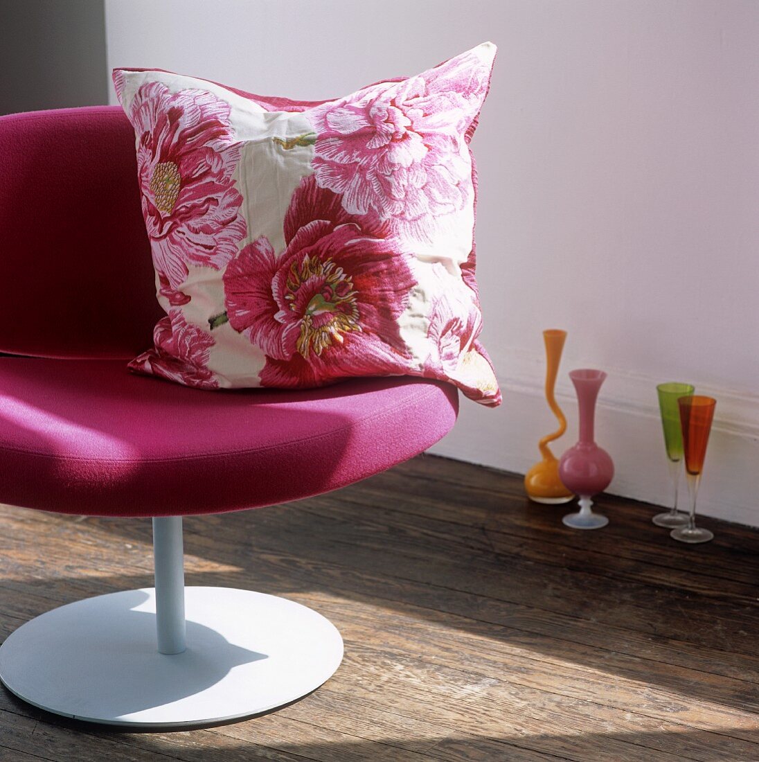 Kissen mit Blumenmuster auf pinkfarbenem Sessel und farbige Vasen und Stielgläser auf Boden