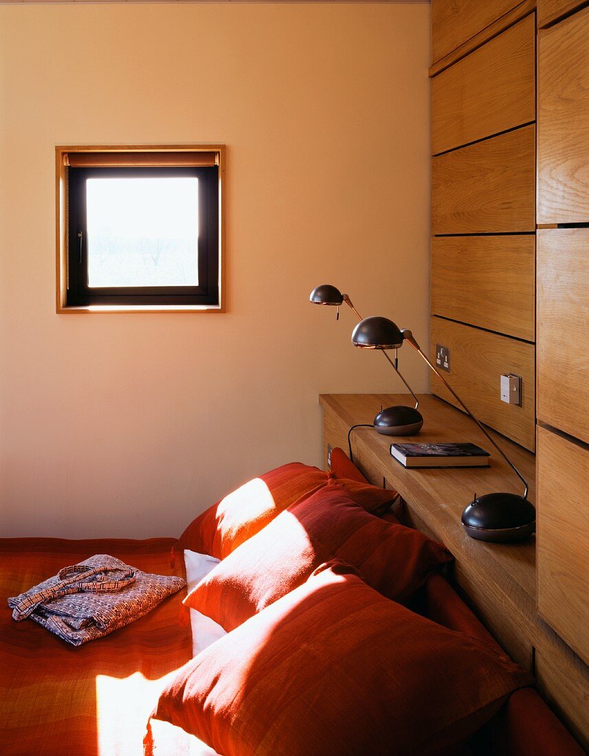 Orange cushions on a bed under a small window and bedside lamps on a wooden shelf
