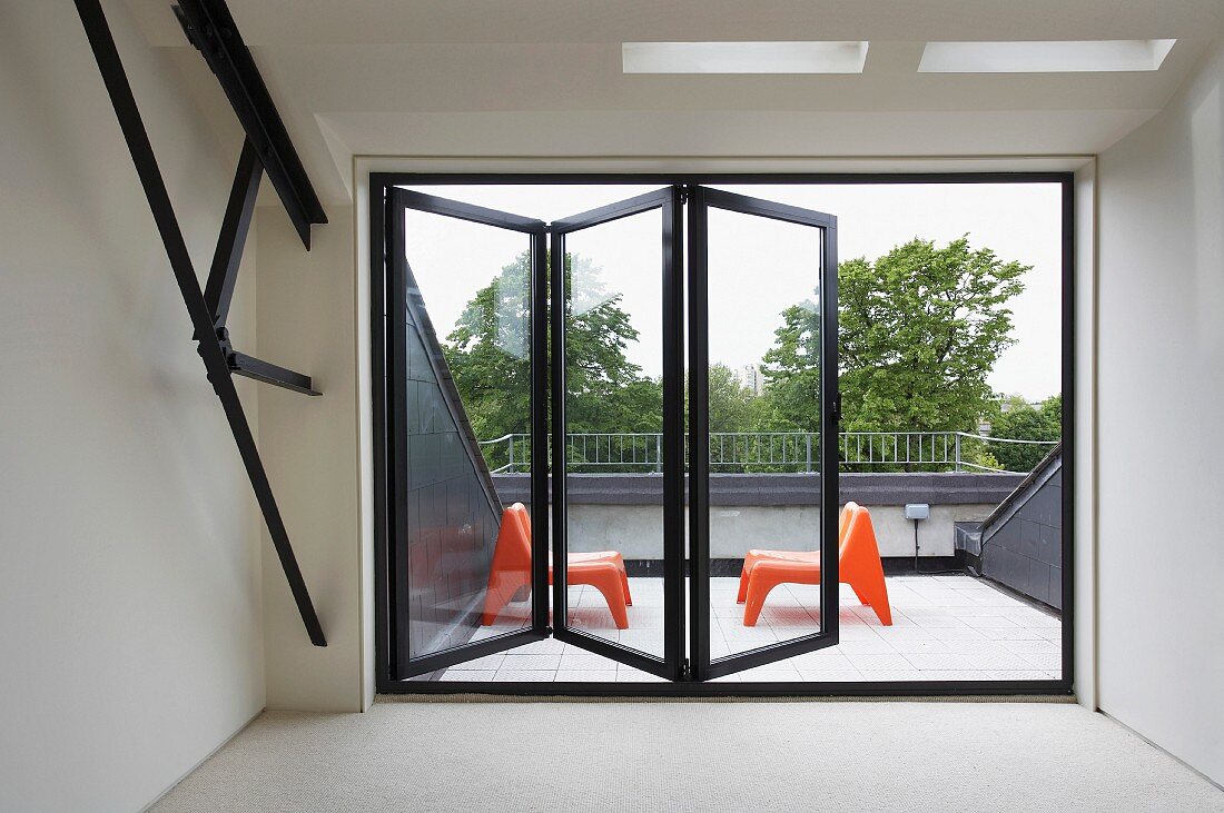 Blick durch halb geöffnete Schiebefaltfenster auf Dachterrasse mit Sesselpaar aus orangefarbenem Kunststoff