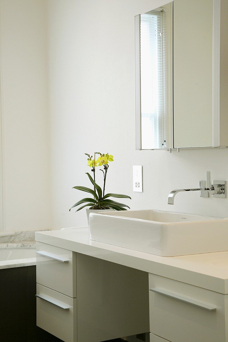 Gelbe Orchidee auf weißem Waschtisch mit eckigem Aufbaubecken und Designer-Wandarmatur unter Spiegelschrank