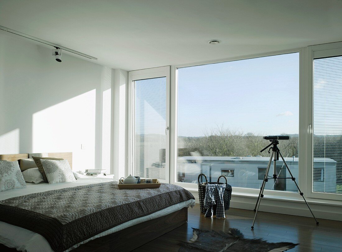 Sonniges Schlafzimmer in Weiß und warmen Grautönen mit Frühstückstablett und Teleskop vor breiter Fensterfront