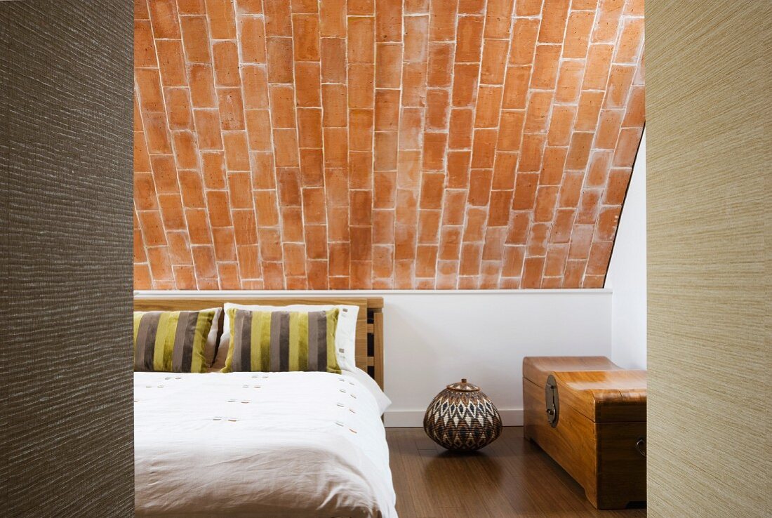 Doppelbett mit gestreiften Zierkissen, edle Holztruhe und Urne mit geometrischen Mustern unter Backsteinschräge