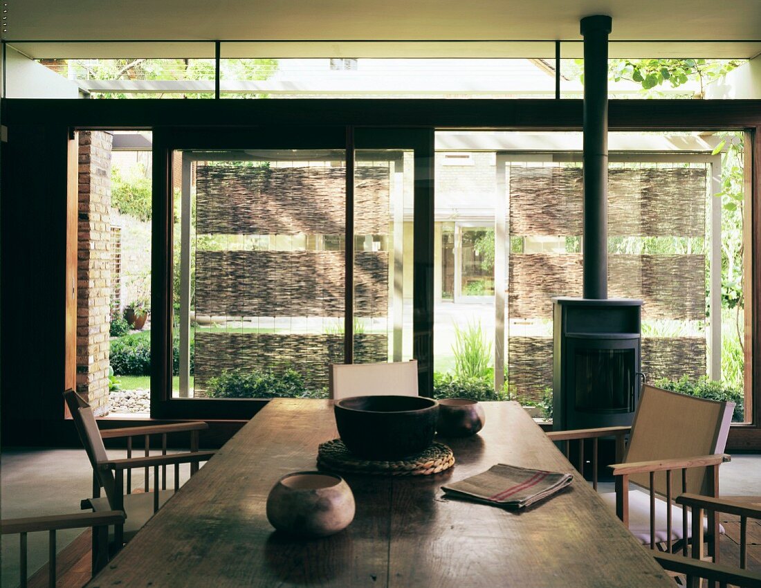 Wohnraum mit Naturmaterialien - antiker Holztisch und Regiestühle vor Fensterfront mit breiter Schiebetür und Sonnenschutzelementen aus Geflecht