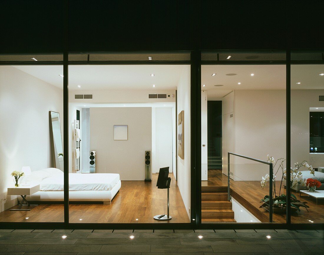 A terrace with floor lights and a view into a modern, illuminated bedroom and a living room