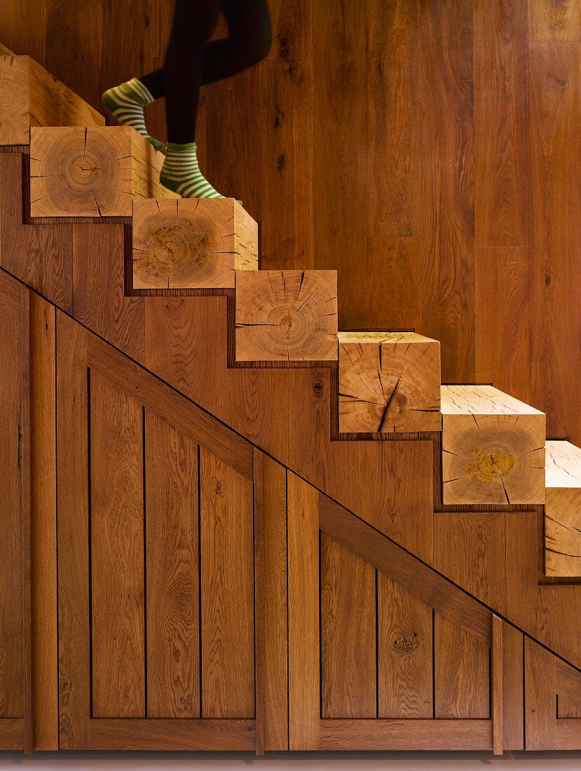 Teilweise sichtbare Person auf offener Holztreppe mit Stufen aus Vierkantholz vor Holzwand