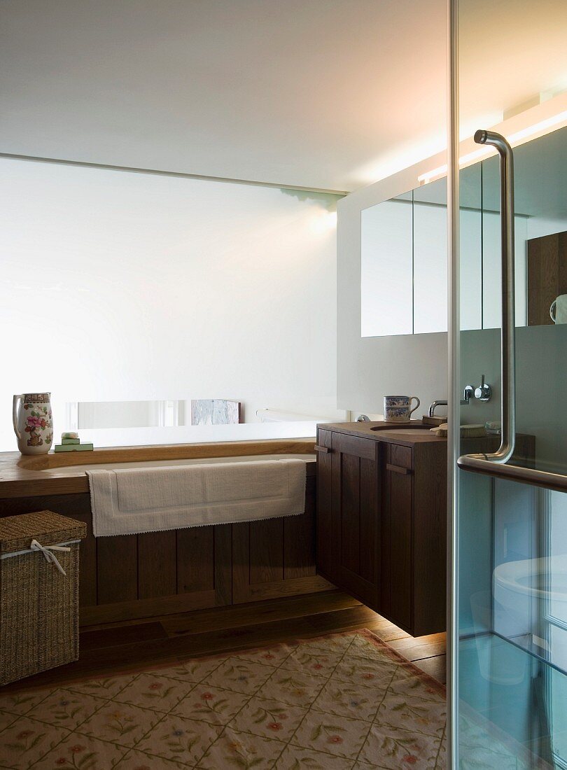 Blick durch offene Glastür auf holzverkleidete Badewanne und Waschtisch im modernen Bad