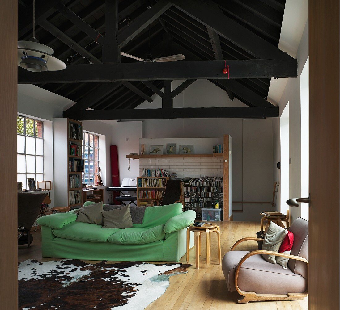 Kuhhaut auf Dielenboden vor grünem Polstersofa im loftartigen Wohnraum mit dunklem Holztragwerk