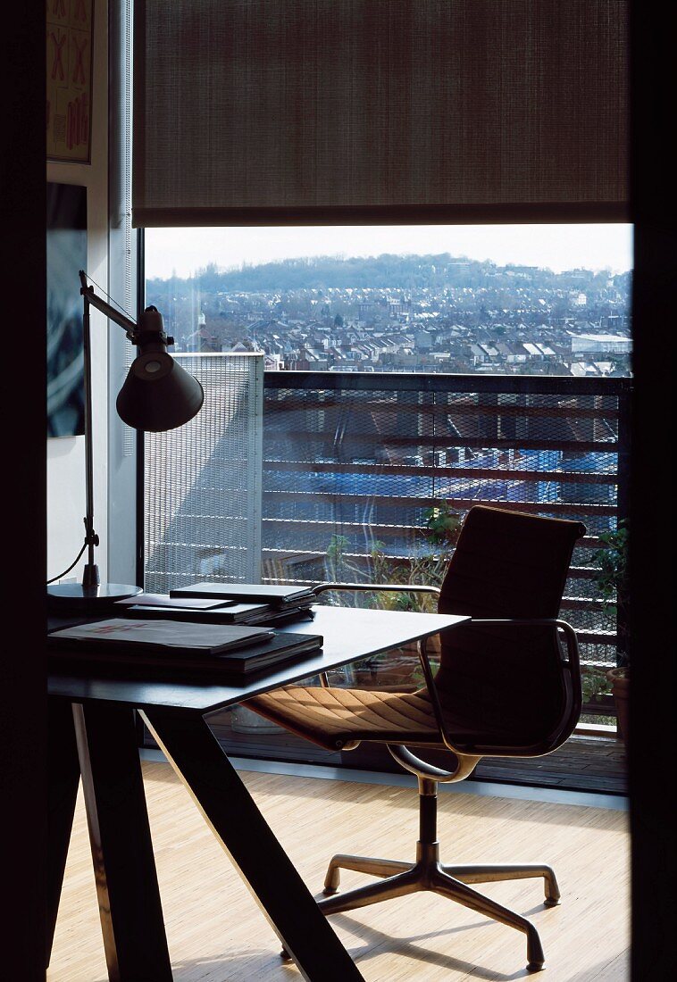 Arbeitstisch mit modernem Bürodrehstuhl vor raumhohem Balkonfenster und Blick auf die Stadt