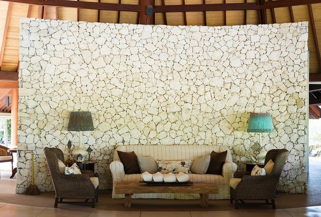 Sofa set and rustic coffee table in front of curved stylised stone partition in tropical hut