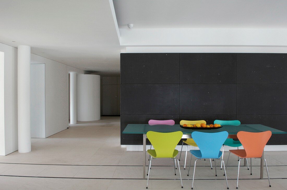 Essplatz mit farbigen 50er Jahre Designerstühlen vor dunkler Trennwand im puristischen Loft; Tischbeine auf Metallschienen im weissen Boden