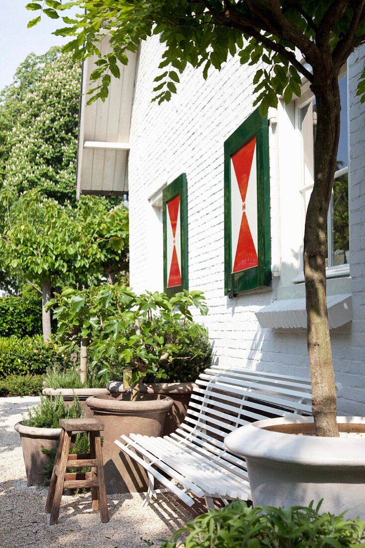 White garden bench and foliage plants in clay pots outside house with white brick facade and shutters painted red, white and green