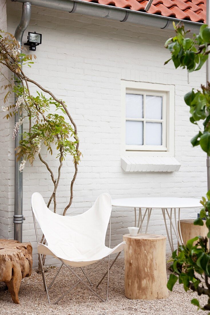 White, 50s-style butterfly chair and tree stump stools in front of white brick house with wisteria
