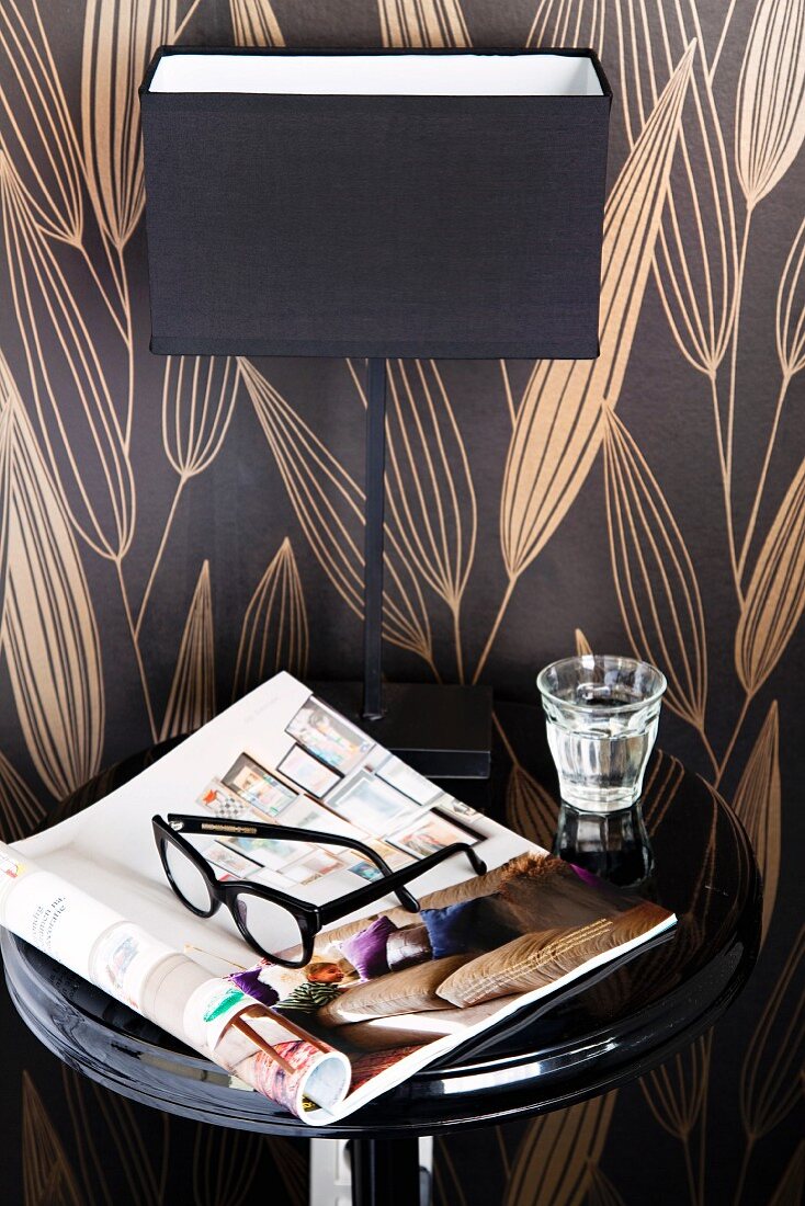 Still-life arrangement against chocolate-brown patterned wallpaper - newspaper, glasses, glass of water and elegant bedside lamp on black lacquered table