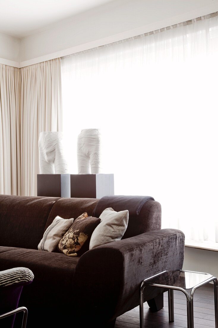 Braunes Samtpolster-Sofa und Stahlrohr-Glastisch vor Fensterfront mit hellen Vorhängen und weissen, halben Torso-Skulpturen