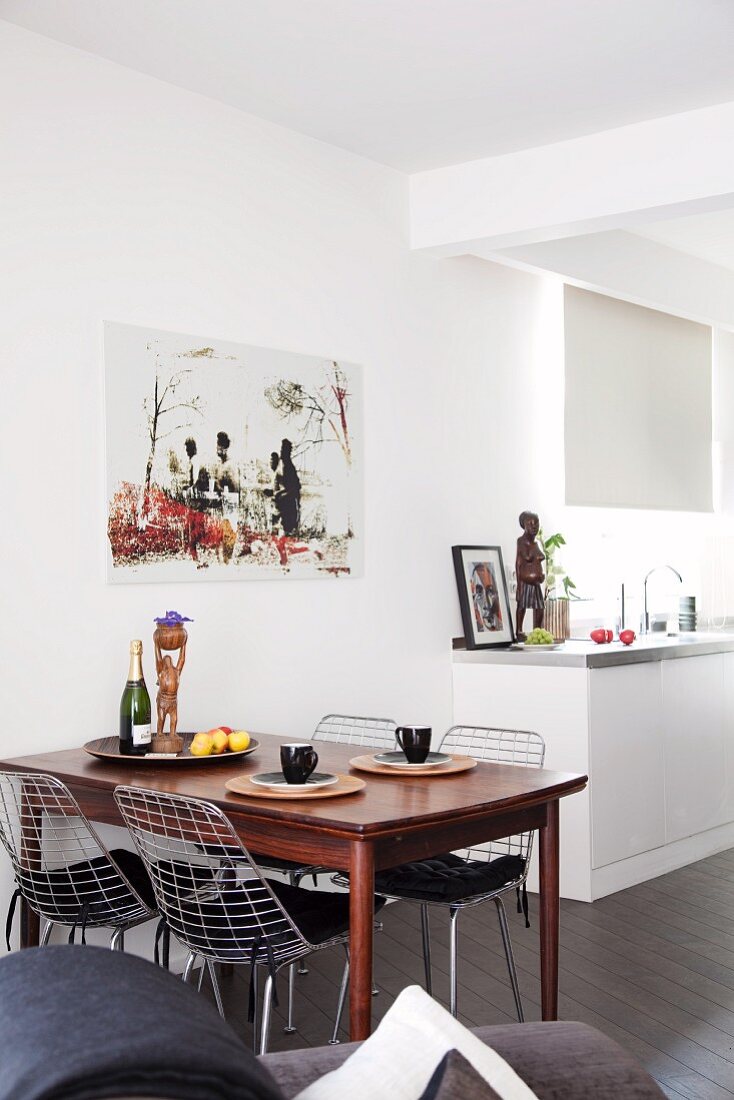 Retro dining table and wire mesh chairs next to white kitchen counter; interior decorated with pictures and sculptures