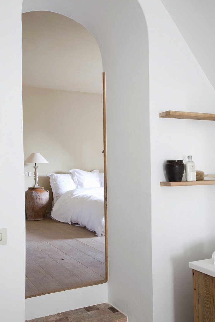 View from bathroom through open, arched door into simple bedroom with terracotta vase used as bedside table