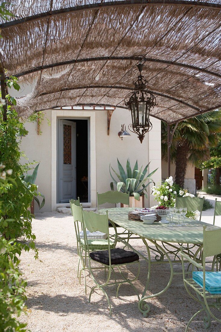 Retro-style metal chairs and table painted pale green below pergola with straw sunshade outside Mediterranean house