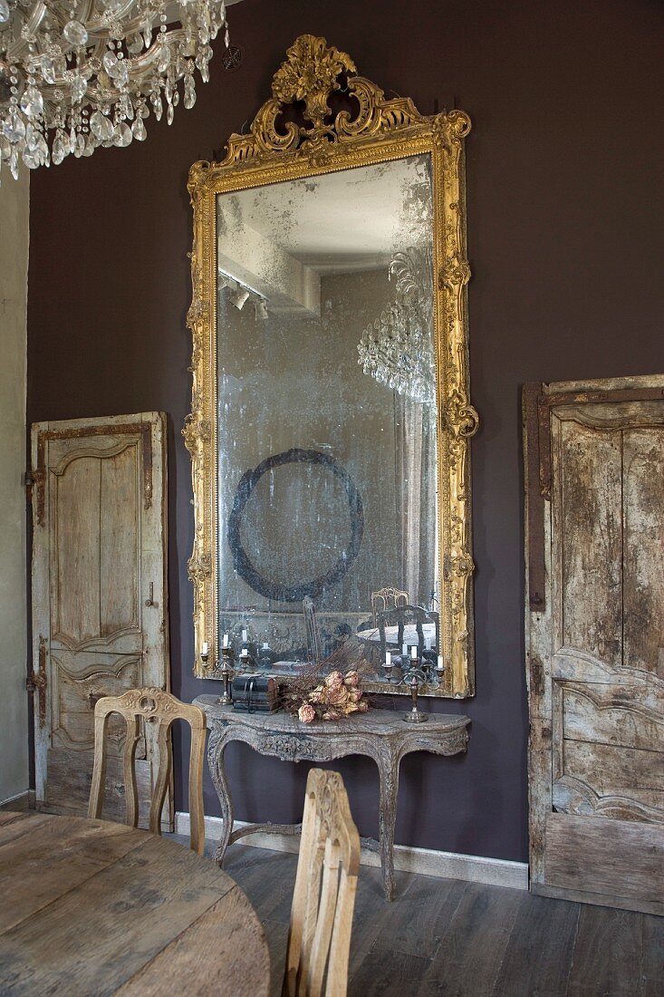 Tall mirror with Baroque gilt frame above Rococo console table against wall painted dark grey flanked by unrestored wooden doors in rustic interior