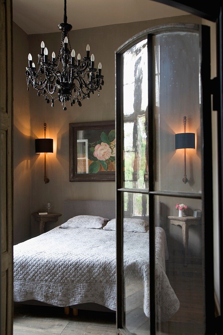 Glass door with metal frame and view of chandelier above bed in simple bedroom with rustic atmosphere