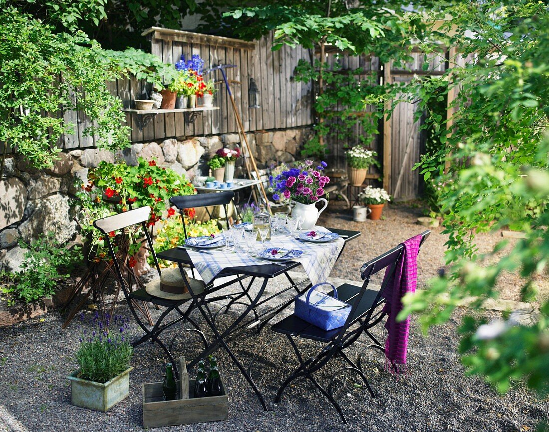 Sonniger, kleiner Garten mit Natursteinmauer, Holzzaun, Blumen und einem gedeckten Tisch auf der Gartenterrasse